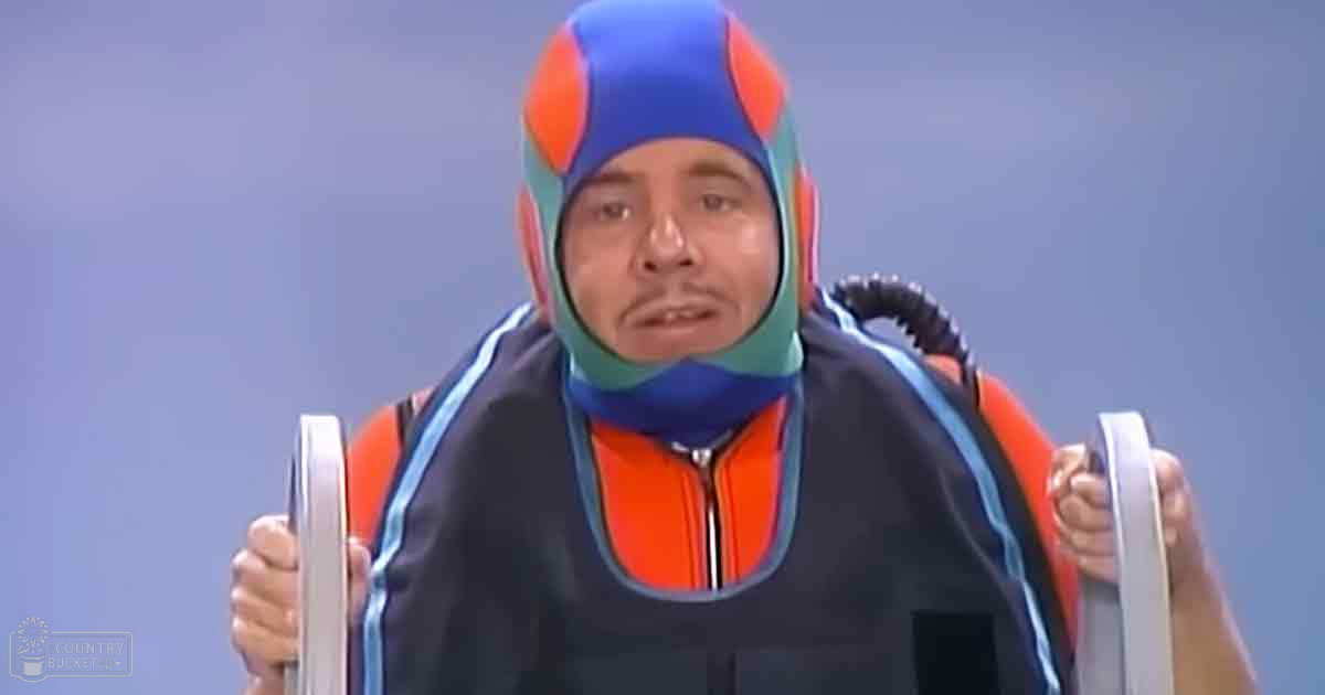 Adult male wearing a tight-fitting orange diving suit with blue accents and geometric patterns, holding metal handles against a plain light blue backdrop under even artificial lighting.