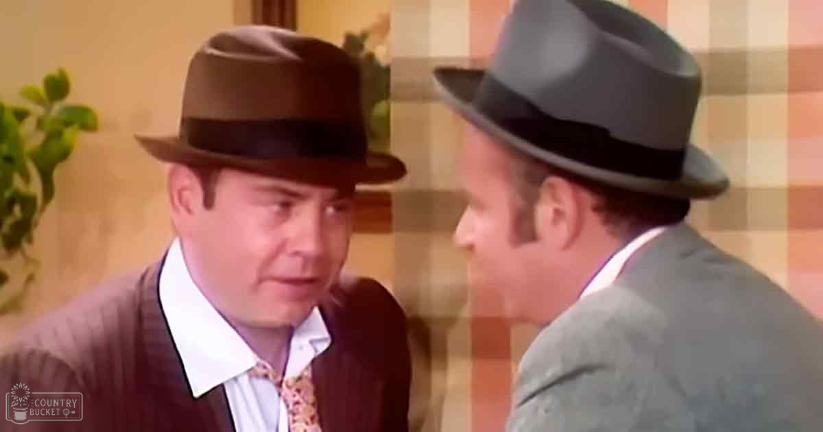 Two middle-aged men wearing fedoras and suits in a casual indoor setting with an orange and beige checkered wall behind them.