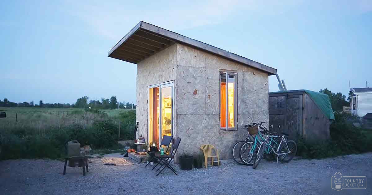 A rural setting with a small house, two folding chairs, a yellow plastic chair, and two bicycles parked outside under a clear blue sky in early evening or late afternoon.
