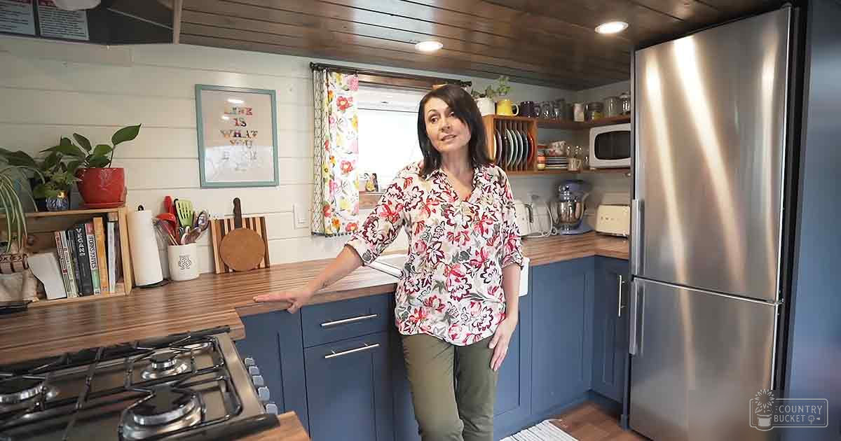 Middle-aged woman wearing a white floral blouse with red and green patterns stands in a cozy kitchen setting, surrounded by wooden countertops and blue cabinets.