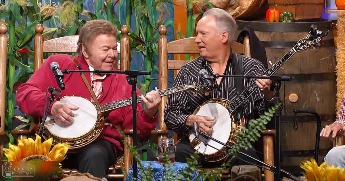Two adult male musicians playing banjos in a studio with farm-themed decorations, suggesting spring or summer season under bright and even lighting.
