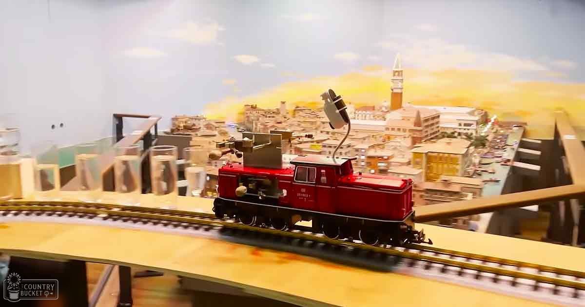 A detailed miniature European-style cityscape backdrop with model train tracks and buildings, likely set during early morning or late afternoon with soft sunlight casting shadows across the structures.