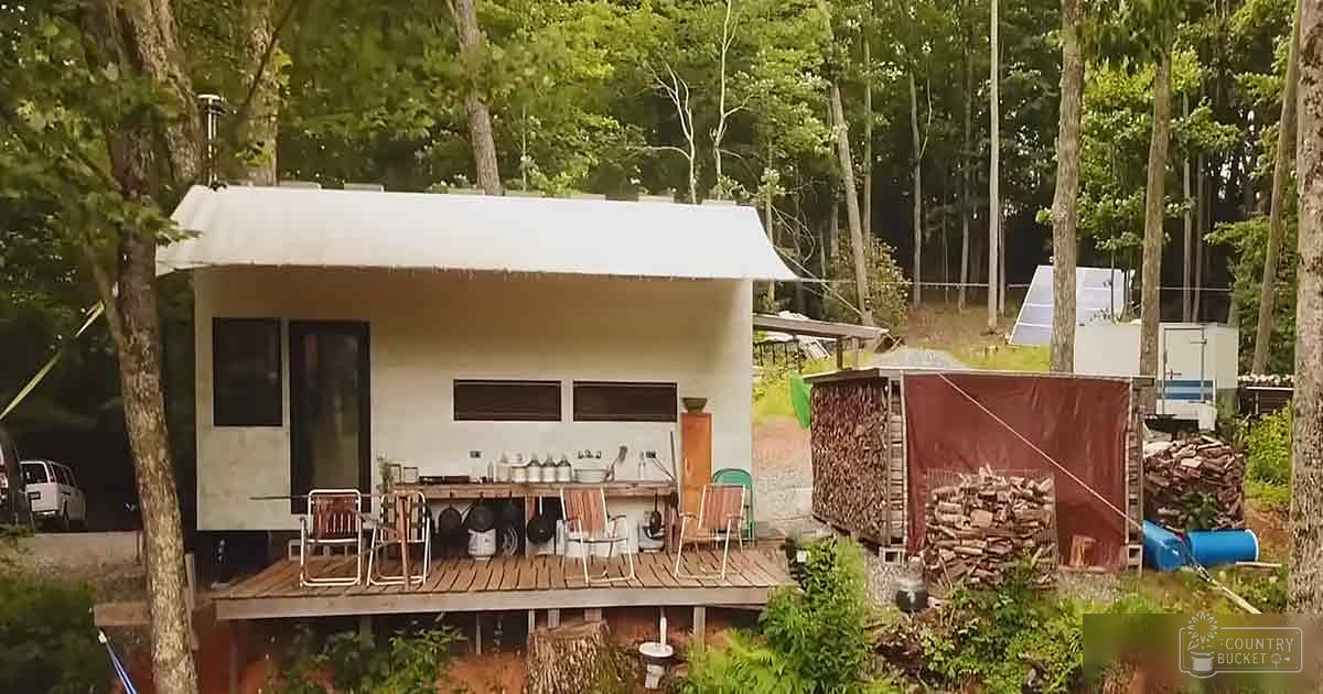 A small white cabin with a wooden deck sits in a forested area, surrounded by trees and various containers on the deck.