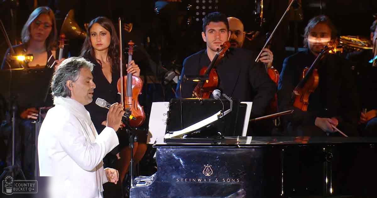 A male performer sings into a microphone on stage with an orchestra, surrounded by darkly lit concert venue elements.