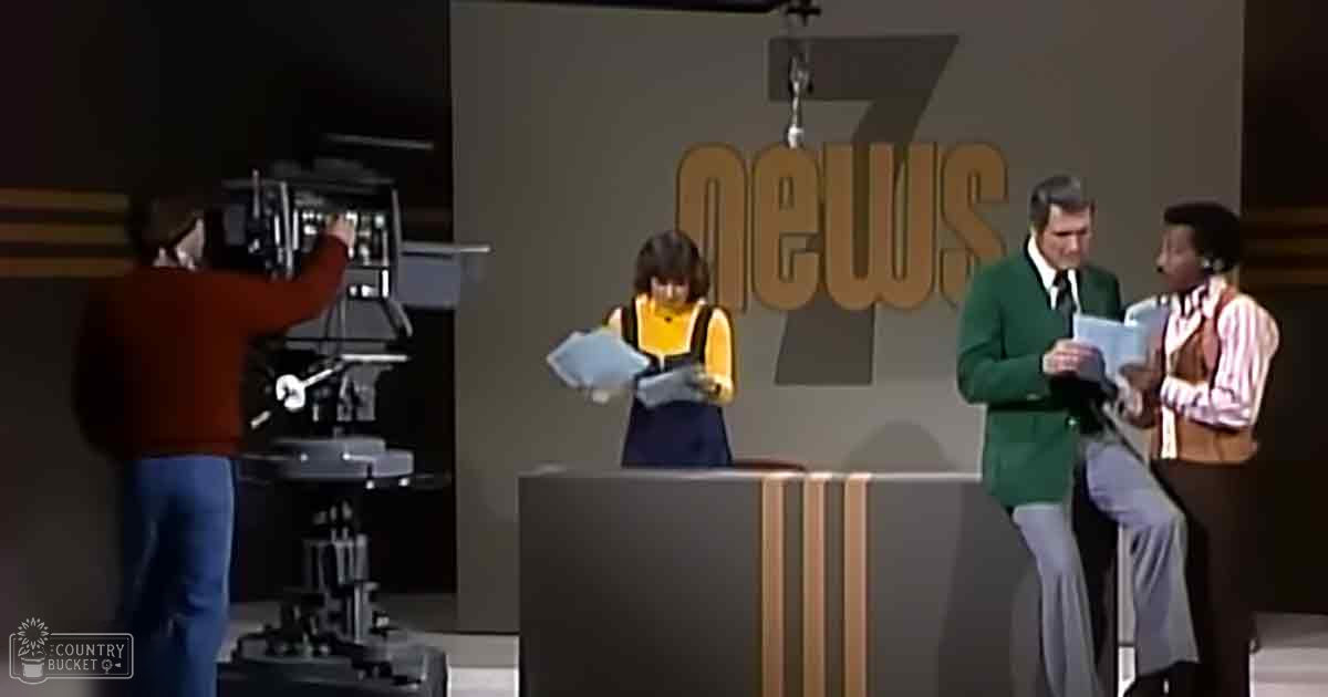 A television studio set with a news desk, large '7 NEWS' sign, camera on a tripod operated by an adult male in an orange sweater and blue jeans. A young adult female in her late teens or early twenties wearing glasses and a yellow top with black trim stands to the left of a middle-aged man in a green blazer over a white shirt with a tie from the mid-20th century, and another adult male holding papers or notes.