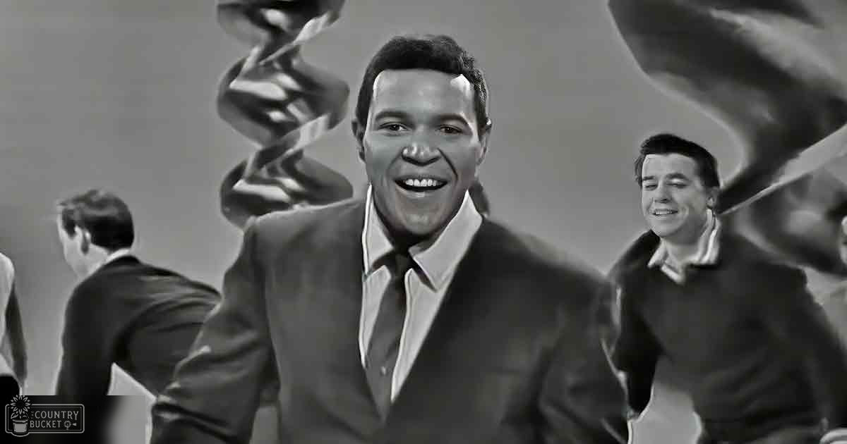 An adult male singer in a dark suit with a young male companion wearing a sweater over a collared shirt indoors against an abstract background.