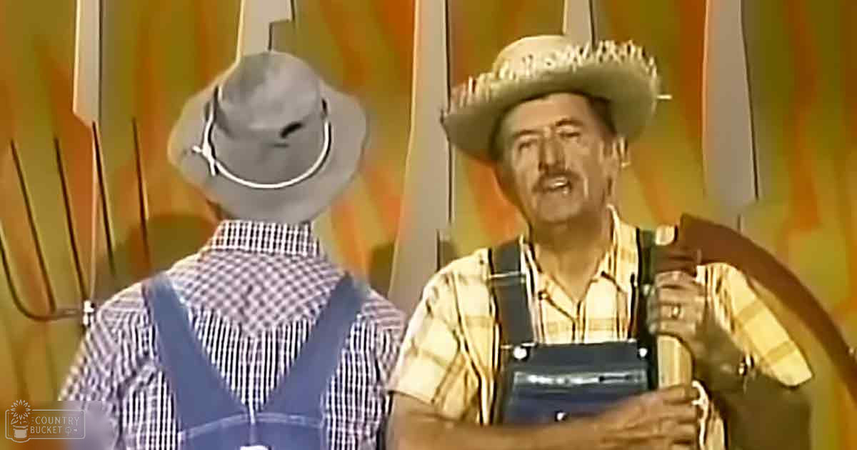 Two adult male performers wearing checkered shirts and suspenders, one holding an accordion, in a rural stage setting with curtains as the backdrop.
