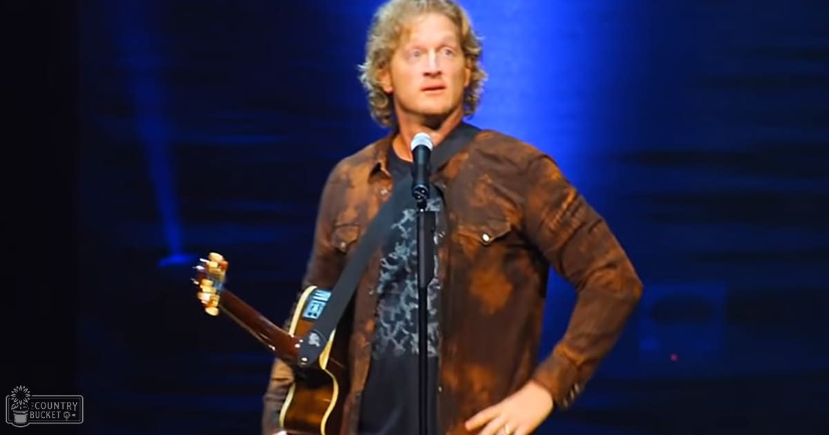 A middle-aged adult male singer wearing a brown patterned jacket over a black t-shirt with a white floral design, holding an acoustic guitar on stage at an indoor venue.