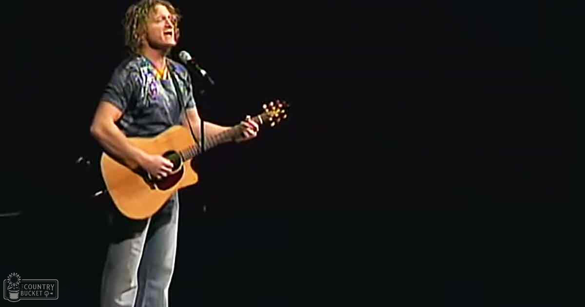 A young adult male performer in a tie-dye shirt with short sleeves, wearing glasses, on an indoor stage at what appears to be a concert venue.