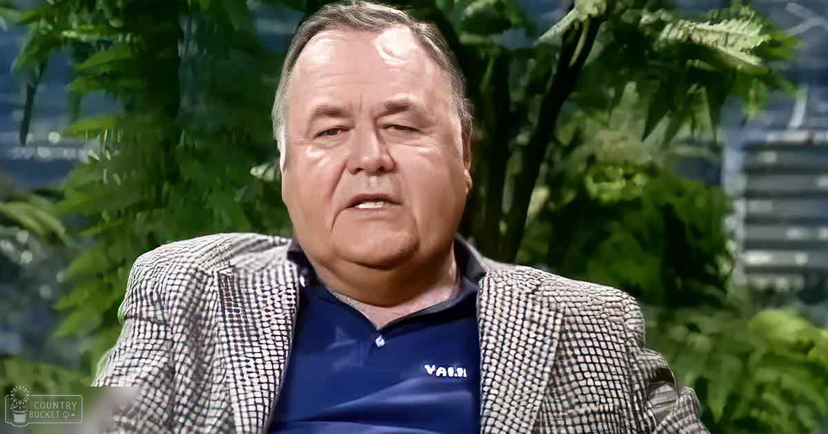 An older adult male in a relaxed fit blazer with a checkered pattern and a blue polo shirt, standing amidst green foliage in what appears to be an outdoor garden or park setting.
