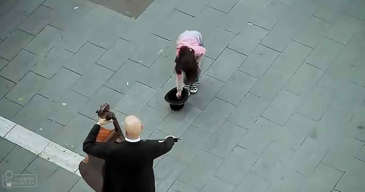 A toddler in a pink hooded jacket and dark pants interacts with an adult male wearing a black suit jacket, white shirt, and brown leather glove outdoors on a paved street.
