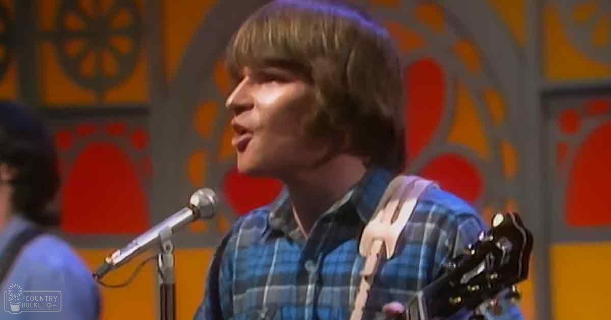 A young adult male performer on stage, wearing a blue plaid shirt, with a child or teen in the background. The setting appears to be an indoor music venue.