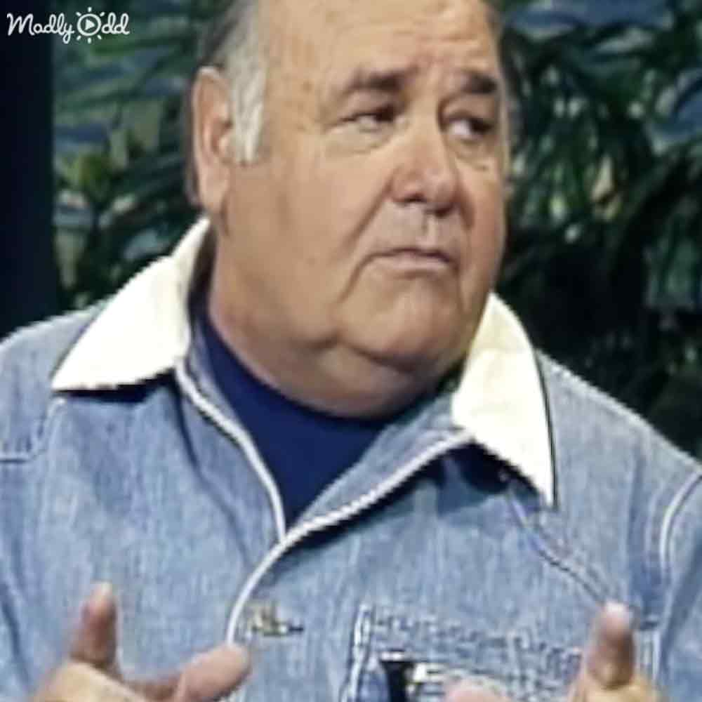 An older adult male wearing a denim jacket with white collar, outdoors amidst greenery under natural daylight.