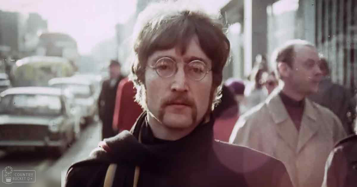 An adult male singer wearing a dark-colored turtleneck sweater stands on an urban street with other pedestrians and a classic car from the late 1960s or early 1970s era.