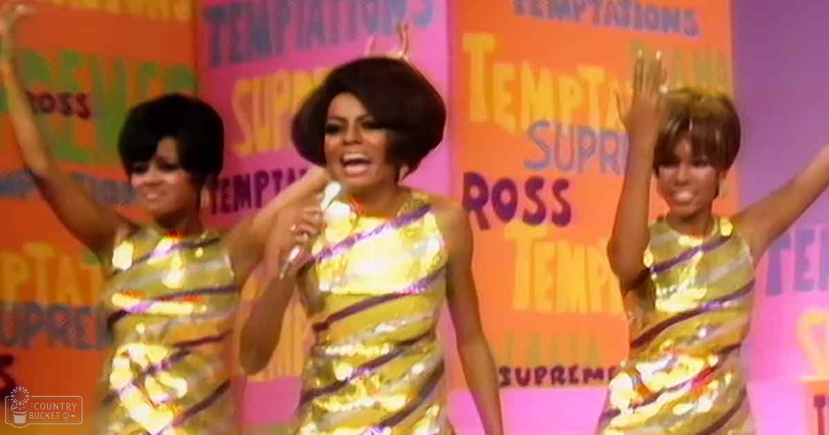 A music concert scene with performers wearing shimmering gold dresses with purple stripes, set against vibrant banners in a stage environment.