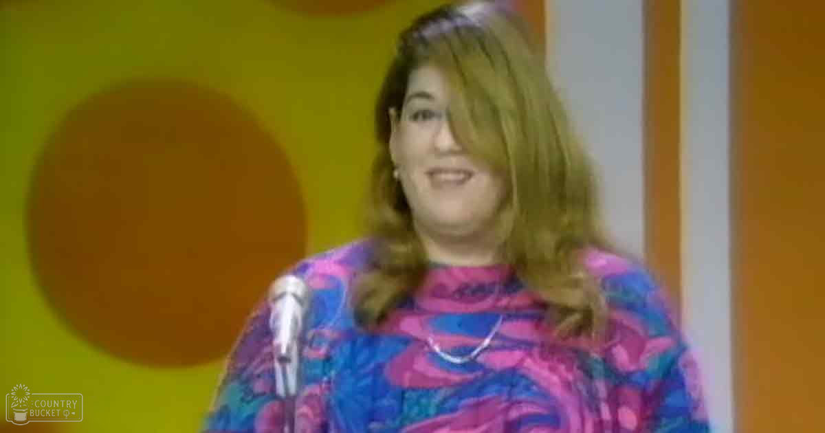 A middle-aged woman wearing a bright pink, blue, and purple patterned top with long sleeves and a round neckline stands indoors near an orange wall with a circular design. A microphone stand is visible on the left side of the frame.