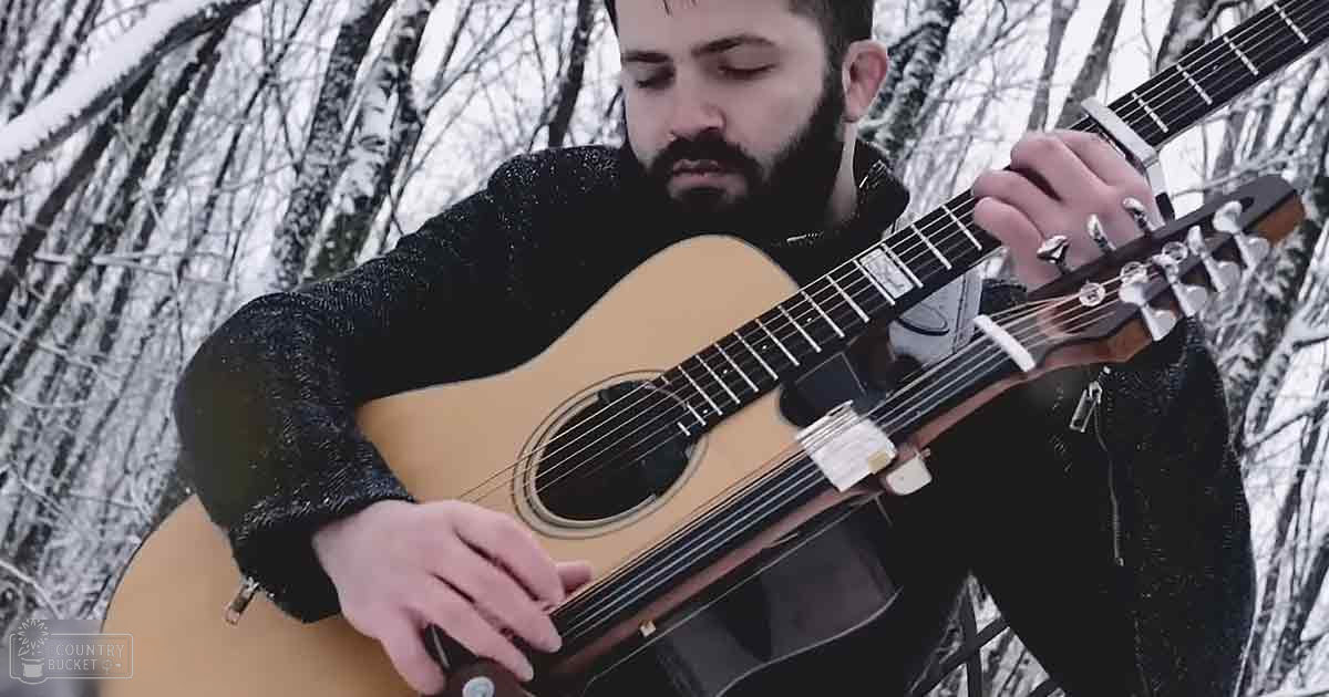 A man in a snowy forest plays an acoustic guitar, wearing a thick black zip-up jacket suitable for cold weather.