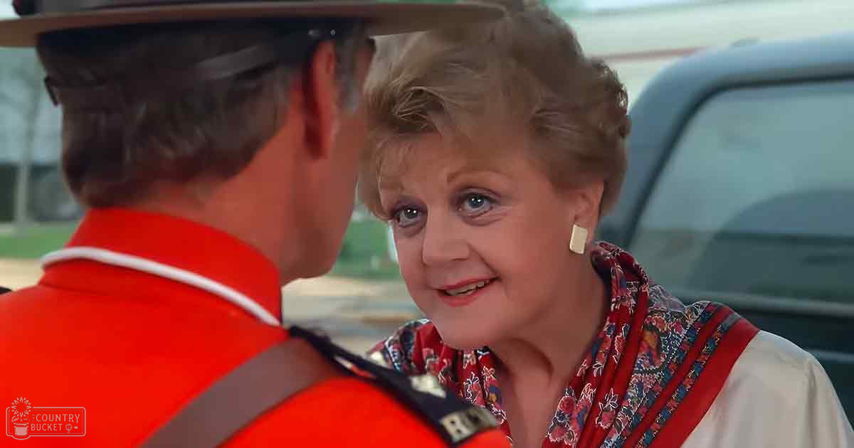 An older adult woman in a white blouse with red and blue floral patterns conversing with another individual wearing an orange safety vest outdoors near part of a vehicle.