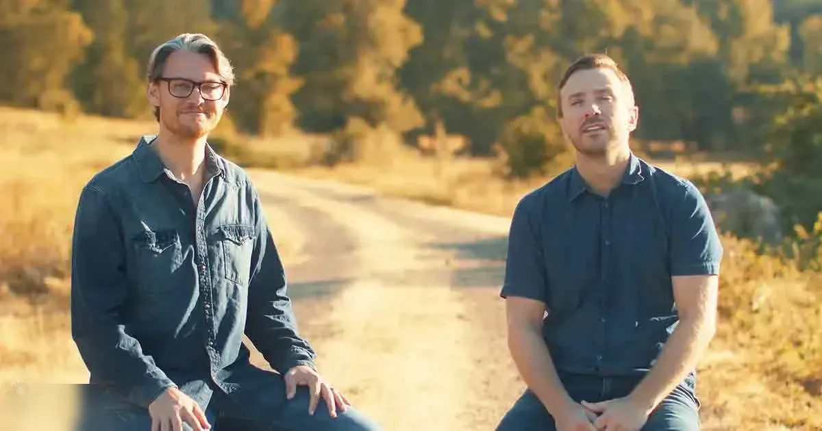 Two young adult males sitting outdoors in a rural setting, wearing casual button-down shirts with collars and chest pockets; the scene suggests early morning or late afternoon lighting.