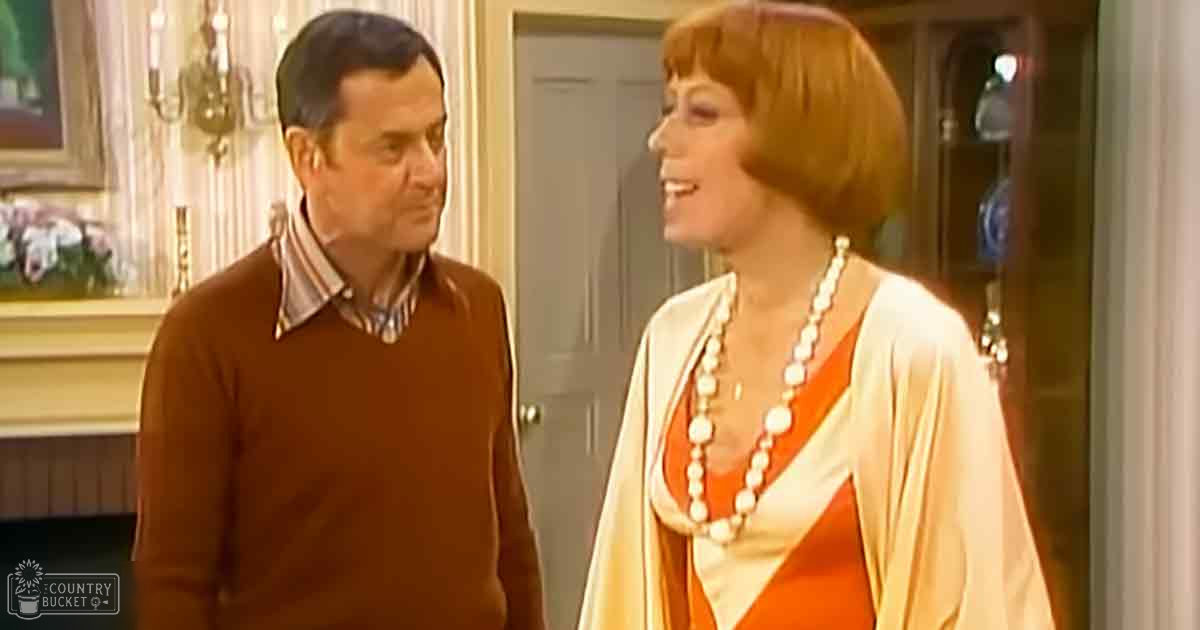 Middle-aged man wearing a brown, relaxed, mid-20th century long-sleeved sweater and middle-aged woman in an elegant orange and white blouse with pearl necklace in a well-decorated indoor setting with natural light during the daytime.