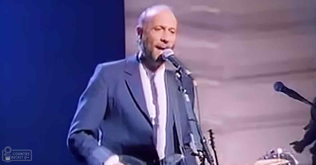 A middle-aged male singer in a light gray suit jacket and white dress shirt performs on stage at an indoor concert venue with a close-up microphone near his mouth.