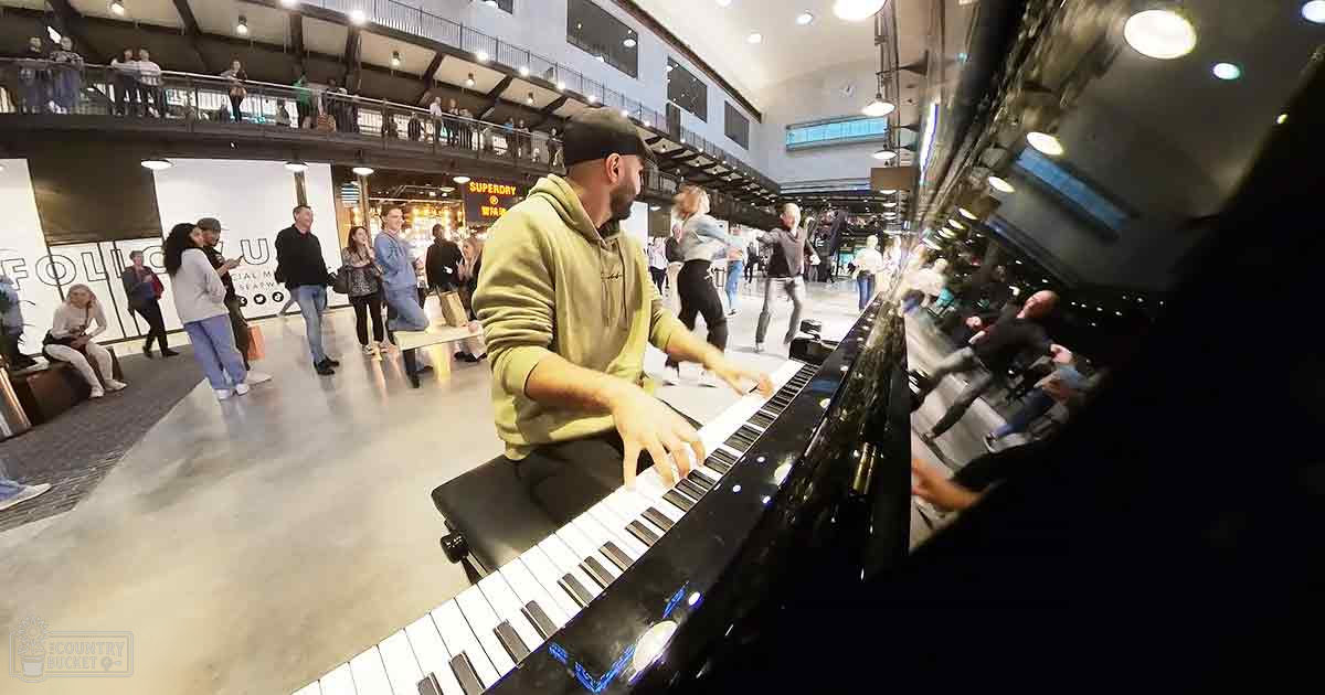 Man with a beard in a green hoodie playing piano, surrounded by people in an indoor market setting; various stalls and displays of goods are visible.