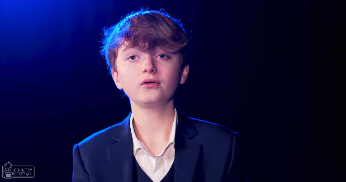 Young adult male in a semi-formal setting, wearing a dark blazer and white collared shirt under dramatic indoor lighting.
