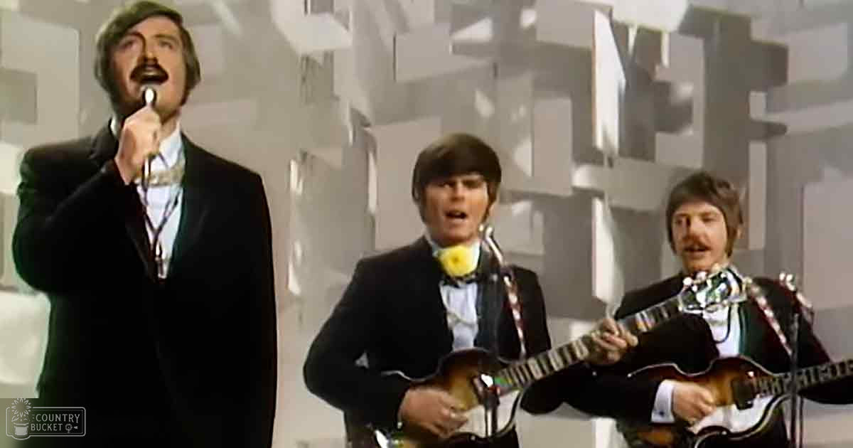 A musical performance scene with three adult male singers in suits on a stage with geometric patterns.
