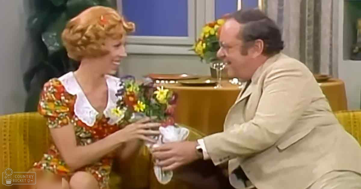 A sitcom scene set in an indoor dining area with two characters, one wearing a floral dress and the other in a beige suit jacket. The setting includes furniture such as a yellow upholstered chair and a table with flowers and wine glasses.