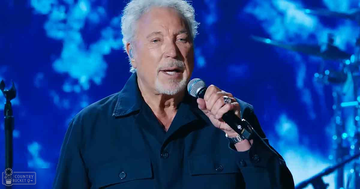 A middle-aged adult male singer performing on a dimly lit indoor concert stage, wearing a relaxed fit dark blue button-up shirt with long sleeves and a collar, holding a microphone.