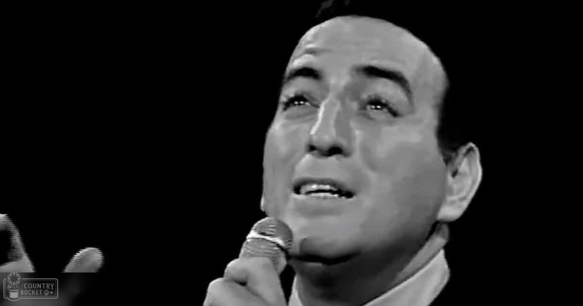 Middle-aged man singing into a microphone in an indoor stage setting with dim lighting, wearing formal attire from mid-20th century style.