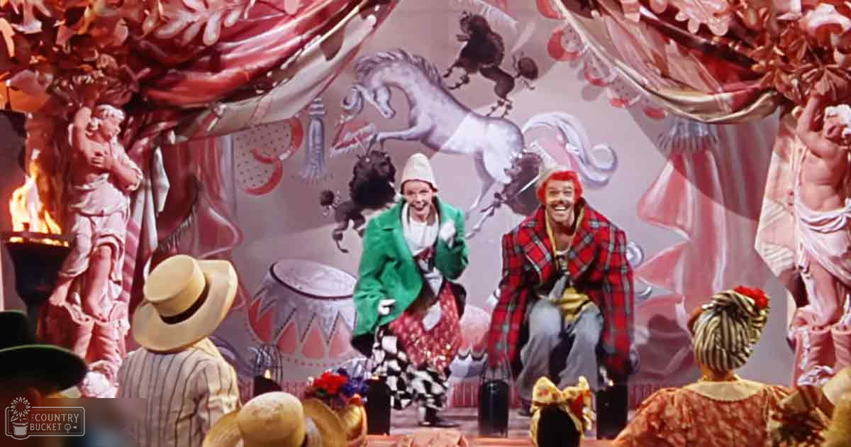A theatrical performance with ornate decorations and classical statues, with two performers in a circus-like setting, surrounded by an audience under bright lighting.