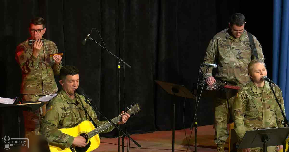 Musical performance by multiple adult performers in camouflage military uniforms indoors with microphones and sheet music, suggesting a live band setting.