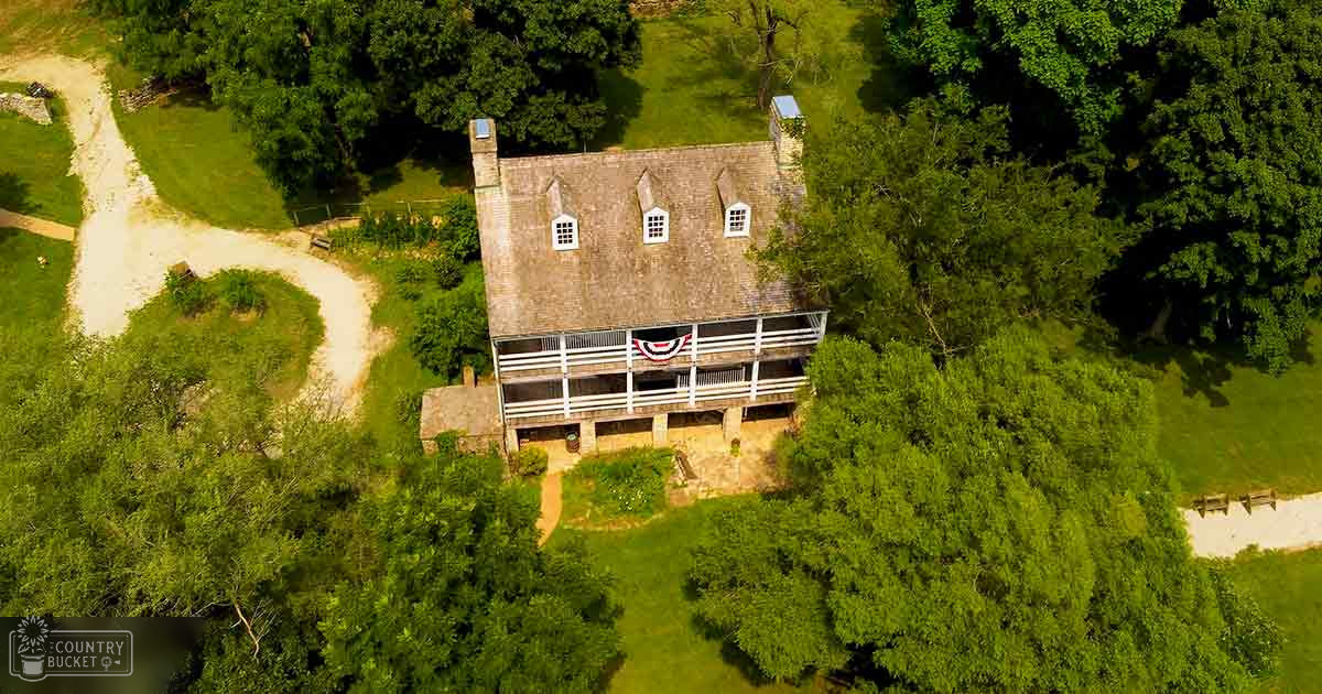 A rural house with a brown roof, white-framed windows, an American flag on its porch, surrounded by trees in daylight.
