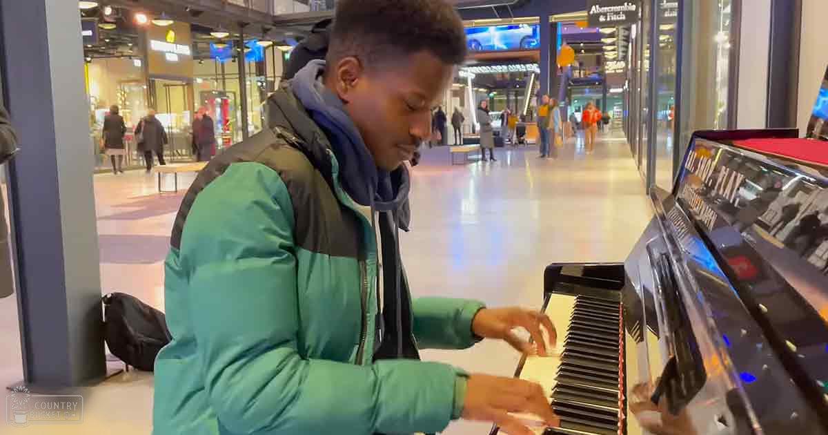 A young adult male wearing a teal and black jacket plays an electronic keyboard in a shopping mall, surrounded by storefronts like Abercrombie & Fitch.