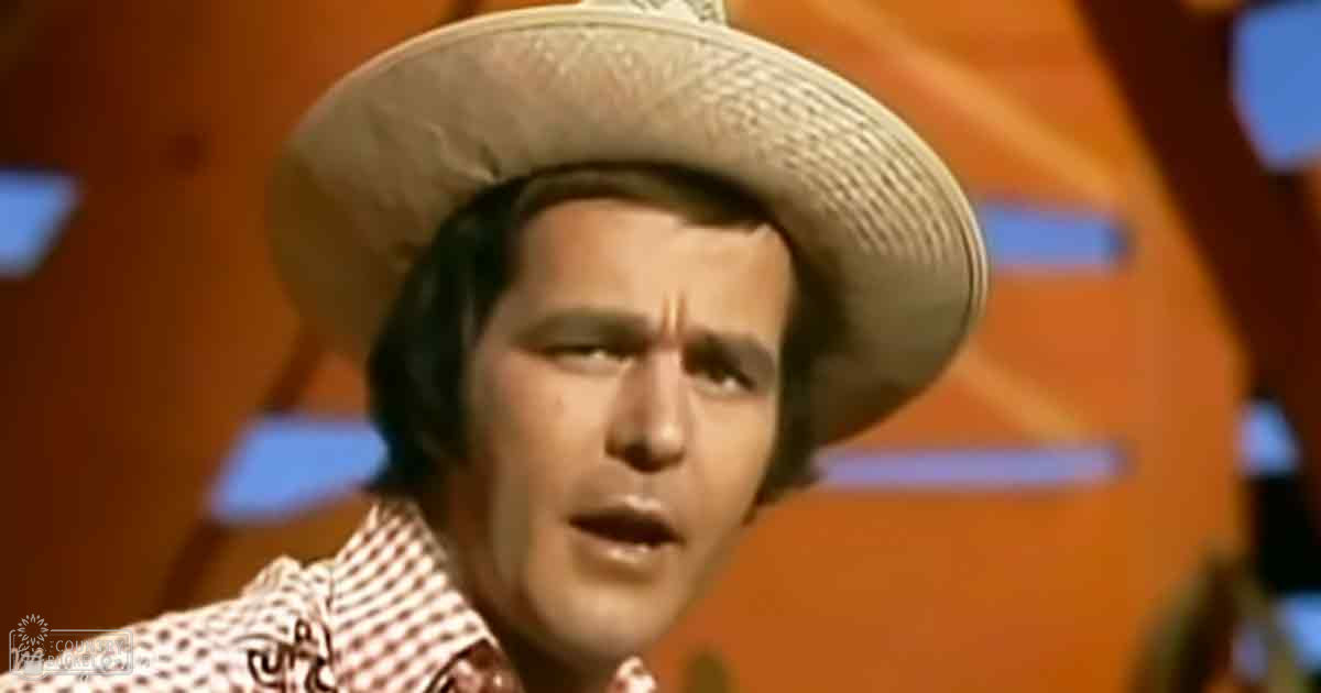 An adult male wearing a red and white checkered collared shirt indoors under warm orange-toned stage lighting, possibly part of a performance setup.