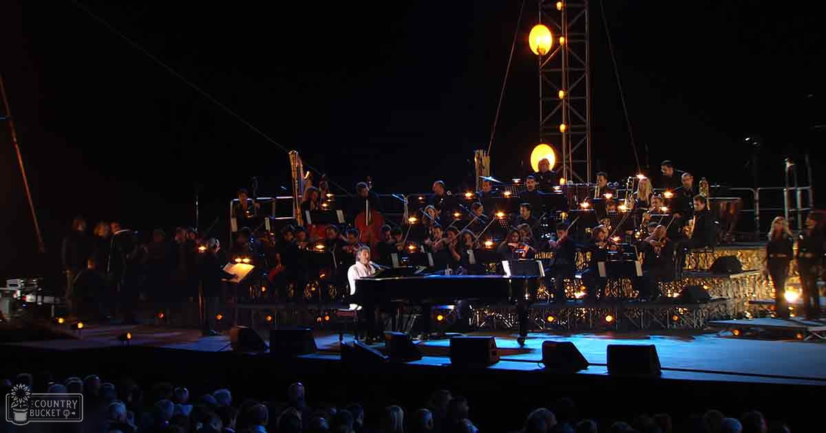 A nighttime outdoor concert scene with an adult male pianist in a dark suit and various ages of orchestra members in formal attire, performing under artificial lighting.