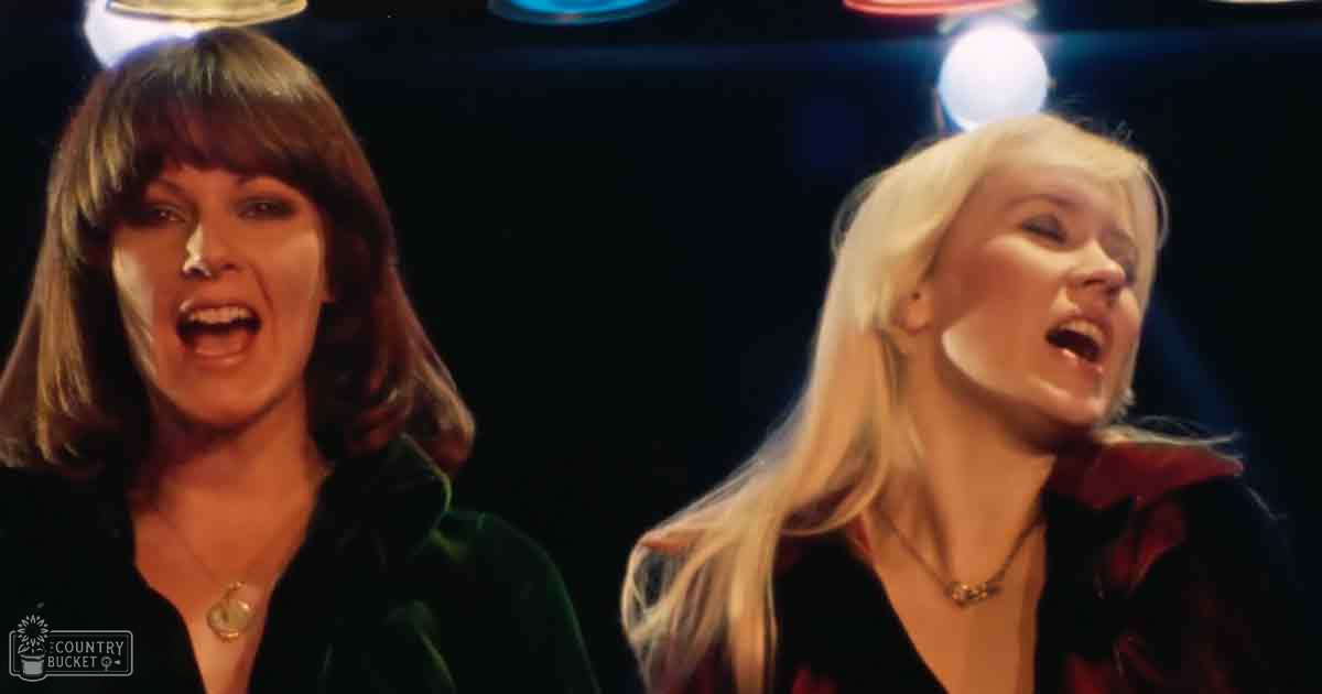 Two women wearing dark velvet jackets with low-cut necklines and pendants, standing indoors under dim lighting that suggests a bar or club setting. The scene is framed by reflections of circular lights in the background.