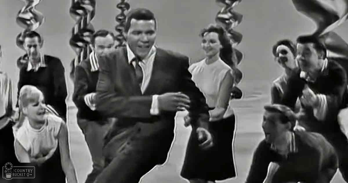 A middle-aged man in a dark suit jacket dances energetically at an indoor event or performance setting with other adults and a toddler in casual attire.