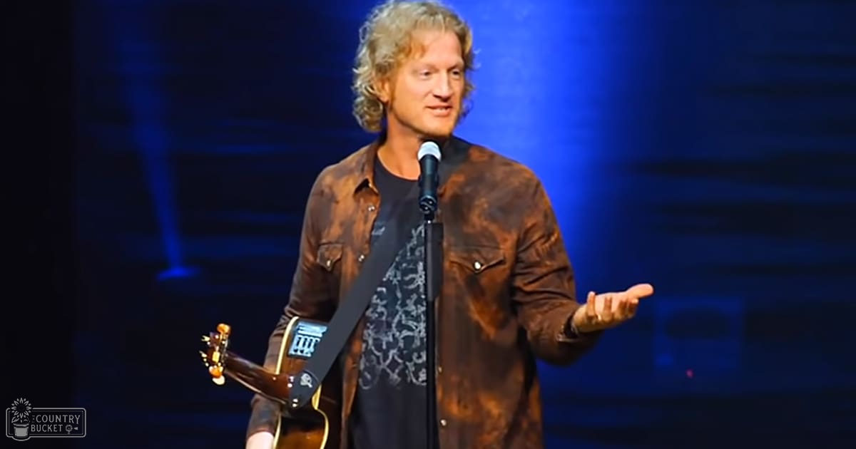 A middle-aged male singer on stage, wearing a brown leather jacket over a black patterned t-shirt with an acoustic guitar and microphone stand visible.
