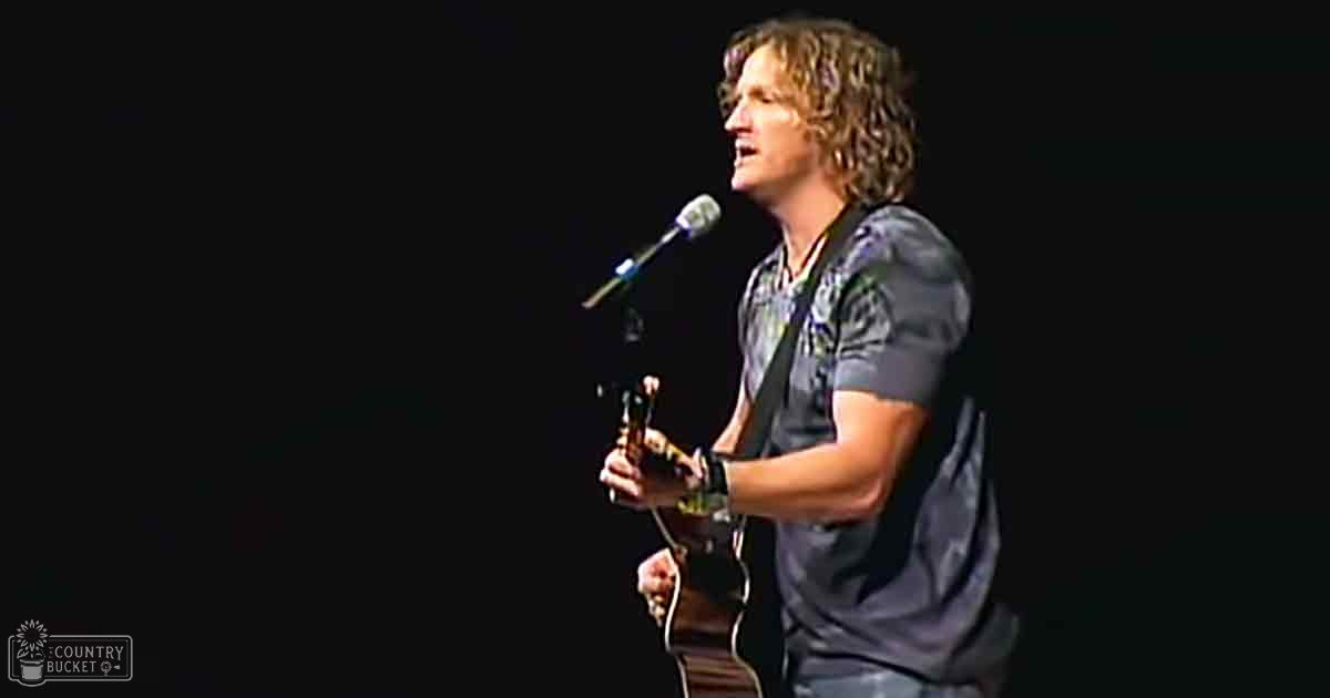 An adult male singer wearing a dark gray, relaxed-fit t-shirt with camouflage or abstract design, performing on stage in an indoor concert venue under low lighting conditions.