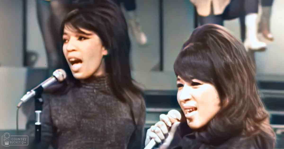Two young adult women singing in a performance setting, wearing dark gray turtleneck sweaters and black low-cut tops with microphones on stage.