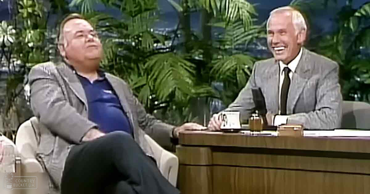 A talk show set with a middle-aged male guest reclining in a chair beside an older adult male seated at a desk; greenery suggests a tropical or garden-like environment, indoor setting with controlled lighting and lush foliage indicating summer or spring season.
