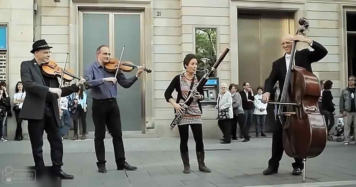 Musicians playing string instruments in an outdoor urban area with a building with large glass doors and windows, surrounded by a casual audience during spring or fall daytime.