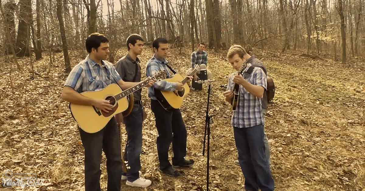 Young adults playing acoustic guitars outdoors in a forested area with dry leaves and trees, possibly autumn or winter; tripod and microphone stand set up for recording music.
