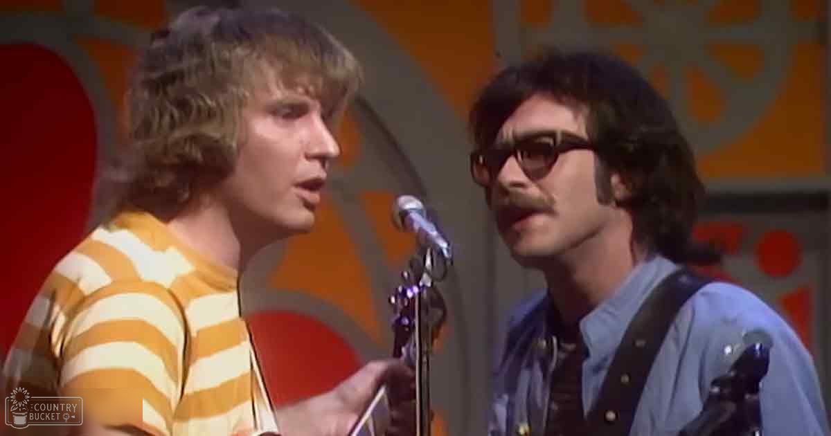 Young adult male singers in casual attire perform against an indoor stage backdrop with orange and red circular patterns, suggesting a 1970s musical performance setting.
