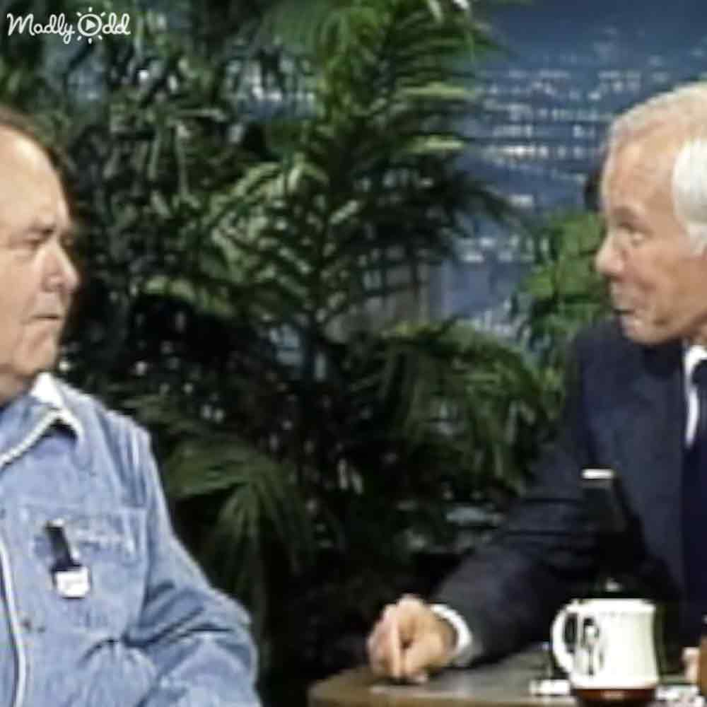 Talk show host wearing a denim jacket with patches, interacting with another older male in formal attire holding a small device; studio setting includes 'Madly Odd' mug and greenery.