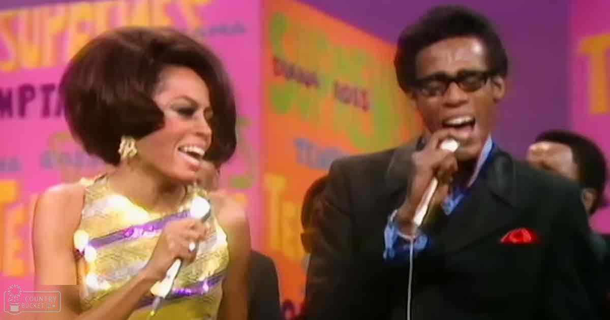 Adult female singer wearing a sleeveless gold dress with purple accents performs on stage, alongside an adult male in a dark suit holding a microphone.