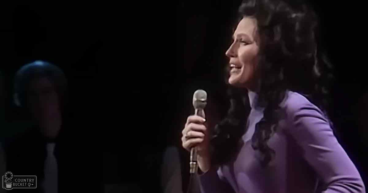 Middle-aged female performer in a purple long-sleeved top, holding a microphone on an indoor stage at night.