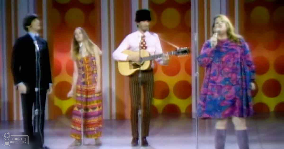 Musical performance with a retro vibe; adult male singers in formal attire and young female performers in flowy dresses, set against an orange and yellow backdrop under artificial studio lighting.